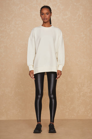 A person with braided hair is wearing an oversized Isabel Ribbed Pullover - Pearl by Noli Yoga, featuring gold side zip details. They have paired it with shiny black leggings and black sneakers. They are standing against a textured, beige background.