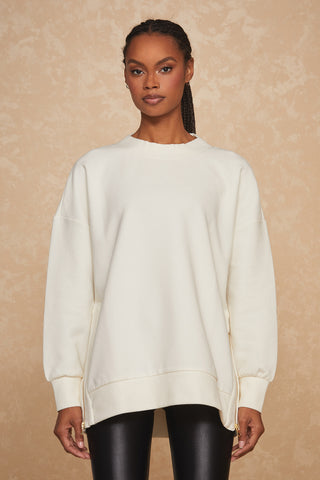 A person with braided hair is wearing the Isabel Pullover in Pearl, an oversized cream-colored crewneck sweatshirt with gold side zips, paired with black leggings. They are standing against a beige, textured background.