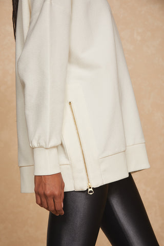 Close-up of a person wearing the Isabel Pullover in Pearl and black leggings. The legging-friendly top features a stylish gold side zip detail near the hem. The background is a neutral beige color.