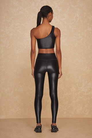 Person with braided hair wearing a black Liquid Curve Bra - Black Gloss and matching black leggings stands facing a beige wall, showcasing the back of the outfit. They are also wearing black athletic shoes.