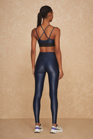 A woman with braided hair, seen from the back, is wearing a navy blue Liquid Jolie Bra with a strappy back and matching high-waisted leggings. The moisture-wicking outfit pairs perfectly with her white and gray athletic shoes. The background is a textured, beige surface.