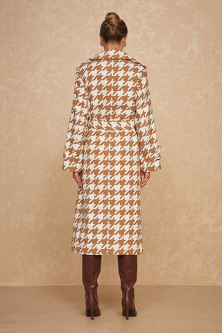 A person with their back facing the camera, wearing a beige and white Juliette Houndstooth Long Coat by Noli Yoga, cinched with a wrap belt. They are also wearing knee-high brown leather boots and have their hair styled in a bun. The background features a textured beige wall.