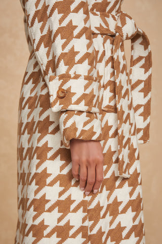 Close-up of a person wearing a beige and white houndstooth-patterned Juliette Houndstooth Long Coat by Noli Yoga. The coat is styled with a wrap belt tied at the waist and cuffed sleeves secured with a wooden button. The person’s hand rests along the side of their body. The background features a light, neutral color.