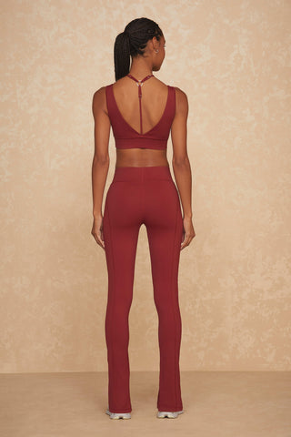 A woman with braided hair stands facing away, embodying chic workout vibes in a maroon two-piece athleisure outfit. She wears a sleeveless crop top with a keyhole back and high-waisted, flared yoga pants, stylishly reminiscent of the Aura Legging - Merlot. White sneakers complete the look against a beige, textured background.