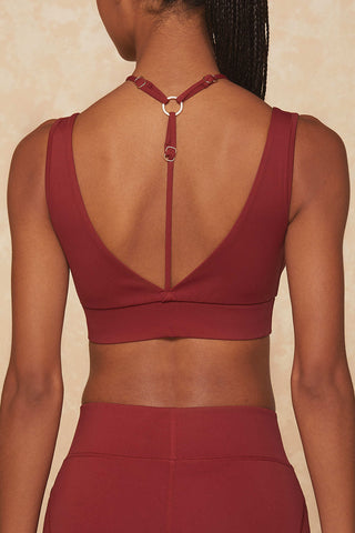 A person wearing the Esme Bra - Merlot and matching leggings is shown from the back. Perfect for low to medium impact workouts, the strappy design features a deep V-neckline and a strap running vertically between the shoulder blades, secured with metallic rings. The background is a light, neutral tone.