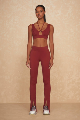 A woman stands against a beige patterned background wearing a chic workout ensemble, featuring an Aura Legging set in Merlot. The outfit includes a sleeveless crop top and high-waisted leggings with front slits at the ankle. She completes the athleisure look with white athletic shoes.