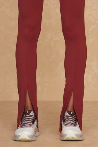 Close-up of a person wearing Merlot-colored Aura Legging with ankle zippers, revealing white and gray athletic shoes underneath. The background is a textured beige wall, perfecting the chic workout look for any athleisure enthusiast.