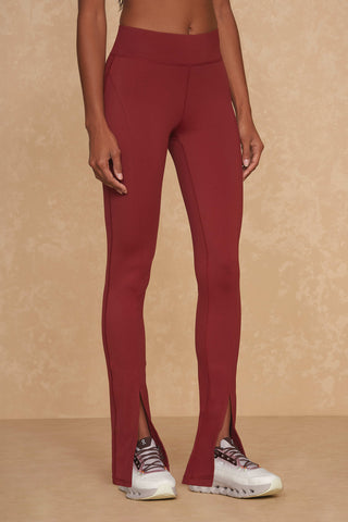 A person wearing maroon high-waisted Aura Legging - Merlot athletic pants by Noli Yoga, featuring slits at the bottom front, and white athletic shoes with black laces, stands against a beige textured background. The person's upper body is not visible in the image.