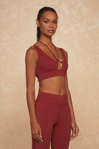 A woman stands against a beige textured background, wearing the Esme Bra - Merlot and matching high-waisted leggings in deep red. Perfect for low to medium impact workouts, the sports bra showcases a unique strappy design at the neckline. She has long braided hair and poses with a relaxed expression.