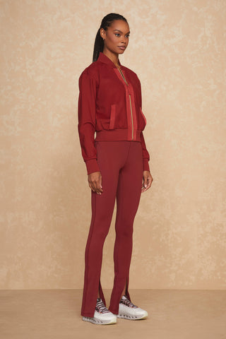 A person stands against a beige textured background, wearing a matching maroon outfit consisting of the Wynn Jacket - Merlot with luxury gold hardware and fitted pants with slight flares at the ankles. They are also wearing grey and white sneakers.