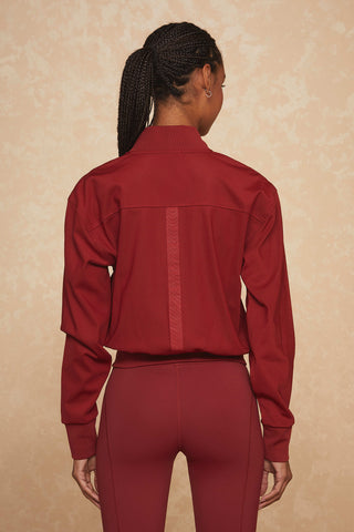 A person with braided hair is seen from the back, wearing a Wynn Jacket - Merlot and matching pants. The background is a light, textured beige color, subtly highlighting the luxury gold hardware on their outfit.