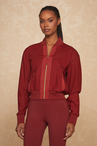 A woman standing against a beige background is wearing a Merlot Wynn Jacket and matching red leggings. The jacket, featuring luxury gold hardware and sheer mesh fabrication, has a high collar and long sleeves. She has long braided hair and is looking directly at the camera with a neutral expression.