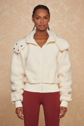 A woman stands against a beige background, wearing the Bianca Sherpa Jacket - Snow from Noli Yoga, characterized by its white fluffiness, oversized silhouette, and high collar. She pairs it with maroon pants. Her hair is styled in neat, short braids and she has a serious expression with her arms hanging by her sides.