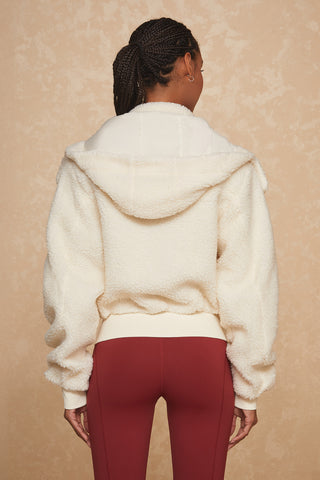 A person with braided hair is facing away from the camera, dressed in the cozy Bianca Sherpa Jacket - Snow by Noli Yoga, which includes a removable hood, paired with dark red leggings. The background features a textured, beige wall.