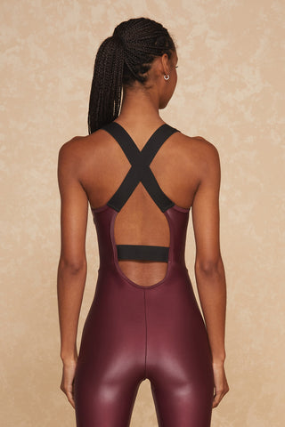 An individual with braided hair is seen from behind wearing the Vixen Jumpsuit in Cherry, which features a sleeveless design, black cross-back straps, and a cut-out detail. The background is a neutral beige with a subtle texture.