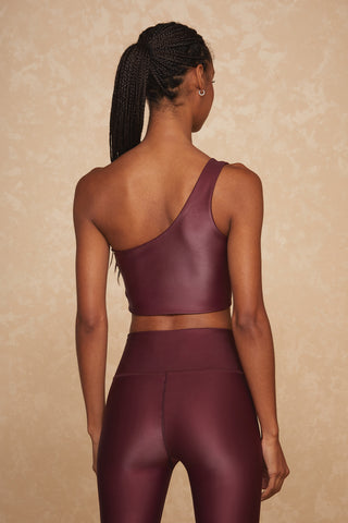 A person with long braided hair is seen from the back wearing the Liquid Curve Bra in Cherry and matching high-waisted leggings. They are standing against a beige, textured background.