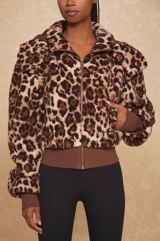 A person wearing the Bianca Sherpa Jacket in leopard print by Noli Yoga with a zip-up front and removable hood stands against a neutral background. The jacket features a large collar, ribbed cuffs, zip pockets, and a fitted waistband. The person is also wearing black leggings.