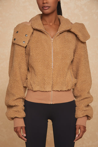 A person is wearing the Bianca Sherpa Jacket - Teddy from Noli Yoga, which features a high collar, zip closure, and ribbed cuffs and hem. The beige jacket includes buttoned straps on the shoulders. The person is also wearing black pants against a beige, textured surface.