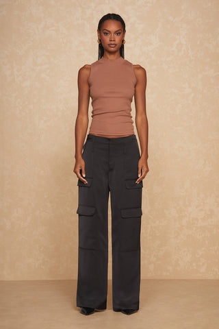 A woman stands against a beige textured background, wearing a sleeveless brown turtleneck and the Milan Pant - Black with wide-leg cargo design. Crafted from satin-like fabric, the mid-rise pants complement her long braided hair as she looks directly at the camera with a neutral expression.