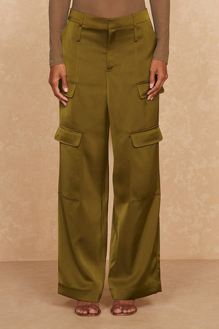 A person is seen from the shoulders down wearing the versatile Noli Yoga Milan Satin Cargo Pant in olive with multiple pockets and a sheer, long-sleeve brown top. The individual stands against a beige textured backdrop, sporting strappy brown sandals.