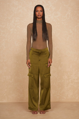 A woman with long braided hair stands against a beige textured wall. She is wearing a sheer, long-sleeve taupe top paired with the Milan Satin Cargo Pant in olive green, featuring large pockets and a high-waisted fit. Made from luxurious silky satin fabric, she looks directly at the camera with a neutral expression.