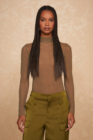 A woman with long braided hair is standing against a textured beige background. She is wearing the sheer, long-sleeved, high-necked Giselle Bodysuit in Olive Mesh along with brown pants that have pockets. She is looking directly at the camera with a neutral expression, showcasing her sleek silhouette.