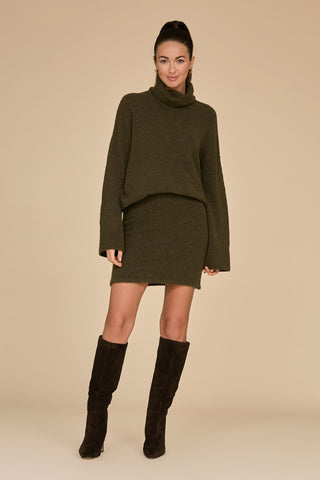 A person wearing the Calista Turtleneck Knit Sweater in pine and knee-high black boots stands against a beige background. The sweater dress features a turtleneck and long sleeves. The person's hair is styled in a high ponytail.