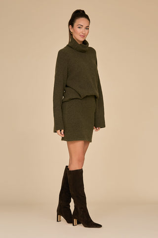 A person wearing a pine-green turtleneck sweater paired with the Calista Knit Skirt - Pine and knee-high brown suede boots stands on a plain beige background, smiling slightly with their arms at their sides.