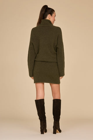 A woman with long hair is standing with her back to the camera, wearing the Calista Knit Skirt in olive green paired with a matching sweater and black knee-high boots against a beige background.