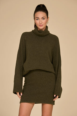 A woman stands against a beige background, wearing the Calista Turtleneck Knit Sweater in pine along with a matching skirt. She has long, dark hair tied back and is smiling slightly.