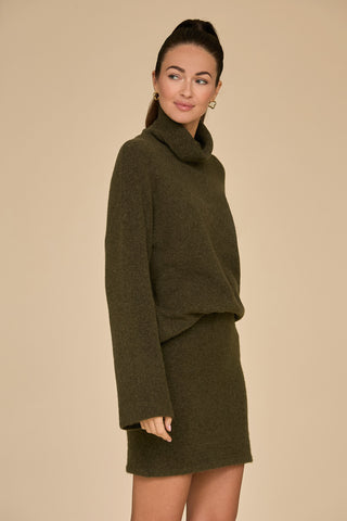 A person poses against a beige background, wearing the Calista Turtleneck Knit Sweater in pine, paired with a matching skirt. They smile slightly while looking to the side, their hair pulled back and adorned with small hoop earrings.