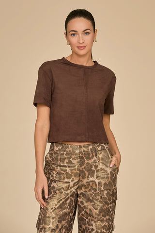 A person dressed in a Naya Suede T-Shirt in Espresso paired with cropped camouflage-patterned pants stands against a beige backdrop. Their hair is styled back, complemented by earrings, and they pose with one hand in their pocket, demonstrating how this shirt works perfectly for layering.