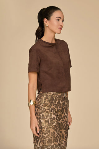 A woman in profile with a ponytail wears the Naya Suede T-Shirt in Espresso, featuring a cropped fit and paired with leopard print pants. She accessorizes with gold hoop earrings and a wide gold bracelet, standing against a plain beige background.