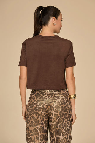 A woman with a ponytail is facing away, highlighting the Naya Suede T-Shirt - Espresso in a cropped fit. Her brown shirt pairs effortlessly with leopard-print pants as she stands against a plain beige background. She wears a gold cuff bracelet on her left wrist, adding an elegant touch to this chic ensemble.