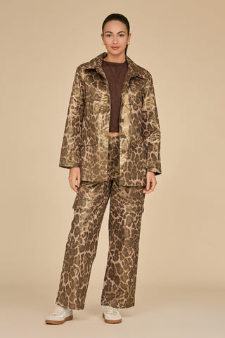 Against a beige backdrop, an individual models a Milan Gold Dusted Cargo Jacket in leopard print, paired with matching pants. Their relaxed pose is complemented by white sneakers. The outfit showcases utility-inspired pockets that seamlessly combine style and functionality.