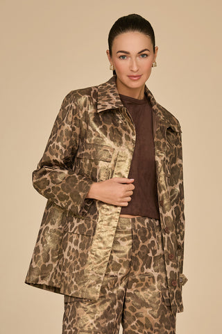 A person poses confidently in the Milan Gold Dusted Cargo Jacket with utility-inspired pockets, featuring a striking leopard print design, paired with matching pants against a neutral background. They sport a dark brown shirt underneath and accessorize with small, elegant earrings. Their hair is styled neatly back, further enhancing the bold ensemble's allure.