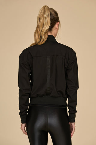 A person with long, wavy hair in a ponytail is depicted from the back, dressed in a Wynn Mesh Jacket - Black, complemented by shiny black leggings. The ensemble, adorned with luxury gold hardware, contrasts strikingly against the plain beige background.