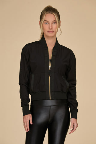 A woman in a sleek Wynn Mesh Jacket - Black, featuring luxury gold hardware, and shiny black leggings stands gracefully against a beige background. Her hair is tied back, with a few strands loose, as she faces the camera directly.