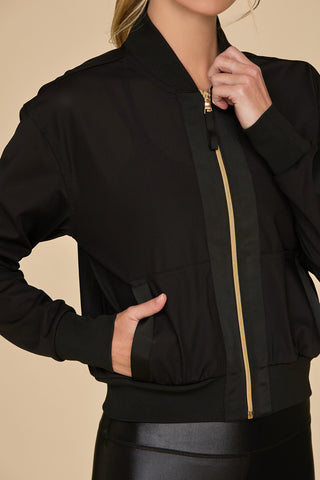 A person wearing a black Wynn Mesh Jacket - Black featuring luxurious gold hardware and black pants. The jacket includes a ribbed collar and pockets. The person's left hand is in one pocket, while the right hand adjusts the zipper. Their head is not visible in the image.