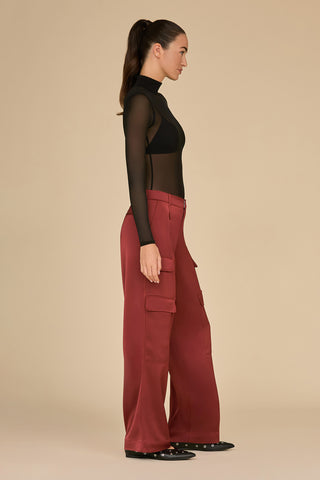 A person stands in profile against a neutral background, wearing a black sheer long-sleeve top and the Milan Satin Cargo Pant in Burgundy, which features wide legs crafted from silky satin fabric and cargo pockets. Their hair is tied back in a ponytail, and they complement the look with sleek black flat shoes.