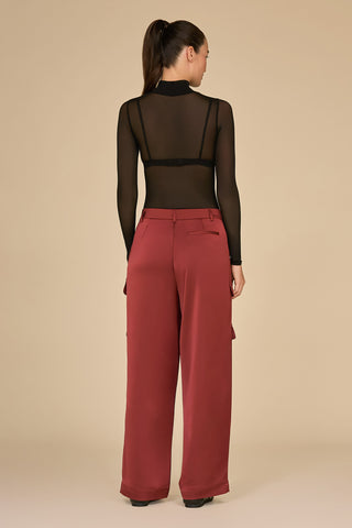 A person with long dark hair tied in a ponytail is shown from behind against a beige background. They are wearing a black sheer long-sleeve top paired with the Milan Satin Cargo Pant in Burgundy, which features a high-waisted and versatile design.