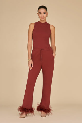 A woman stands against a plain background, wearing the Talia Ribbed Tank in Burgundy, paired with matching pants with feathered cuffs. The ensemble highlights her slim silhouette, complemented by nude heels and a gold bracelet. Her hands rest casually in her pockets, adding effortless charm.