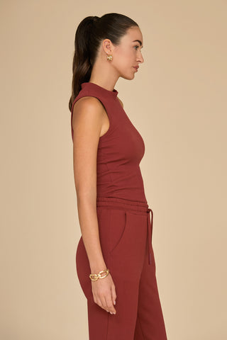 A person with long hair tied back is wearing the Talia Ribbed Tank in burgundy, facing sideways against a plain beige background. Their sleeveless outfit features an ultra-soft rib and is accessorized with a bracelet and earrings, highlighting a slim silhouette.