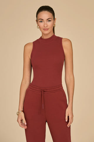 A person with long hair is wearing the Talia Ribbed Tank in burgundy along with matching pants, accentuating a slim silhouette. The ultra-soft rib material enhances their neutral expression against a plain beige background as they stand facing the camera.