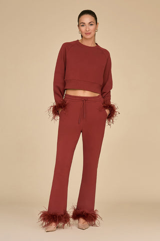 A person is wearing a matching brown French Terry loungewear set with ostrich feather trim on the cuffs and ankles, standing against a neutral background. Their hair is tied back, and they're sporting beige high heels that perfectly complement their chic Rada Feather Trim Pant in Burgundy.