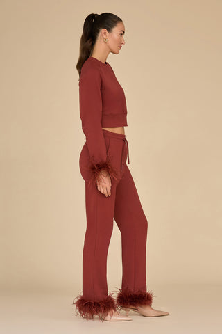 A woman in a burgundy matching set, featuring the Rada Feather Trim Pant - Burgundy, stands in profile against a beige background. The French Terry outfit includes a crop top and pants with ostrich feather trim on the cuffs. She has long hair tied back in a ponytail and is paired with beige shoes.