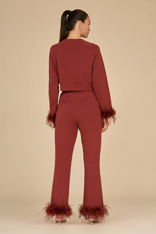 A woman stands turned away, displaying a fashionable ensemble featuring the Rada Feather Trim Pant in Burgundy and a coordinating maroon French Terry top. Both pieces are elegantly accentuated with an ostrich feather trim at the cuffs. Her hair is tied back in a ponytail against a neutral beige backdrop.