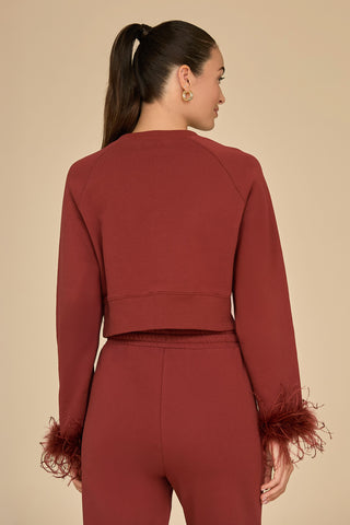 A woman with a ponytail stands with her back to the camera, dressed in a burgundy Rada Feather Trim Crewneck made from French Terry fabric. The long-sleeved top and pants are adorned with ostrich feather trim on the sleeves, set against a plain beige background.