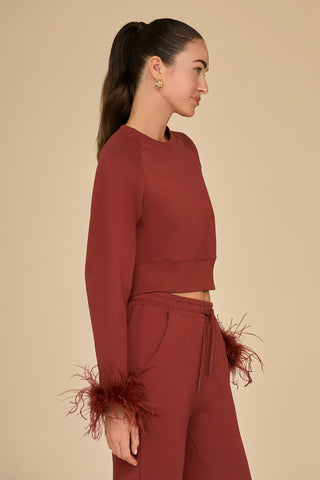 A woman with long dark hair in a ponytail is wearing the Rada Feather Trim Crewneck in burgundy, featuring ostrich feather trim on the cuffs. She stands sideways against a beige background, accessorized with small hoop earrings.