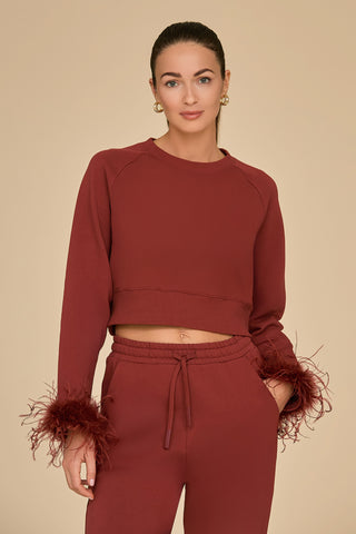 A person dressed in a burgundy matching set, featuring the Rada Feather Trim Crewneck adorned with ostrich feather cuffs and drawstring pants made from French Terry fabric, stands against a beige background. They have short hair and are looking directly at the camera with a neutral expression.